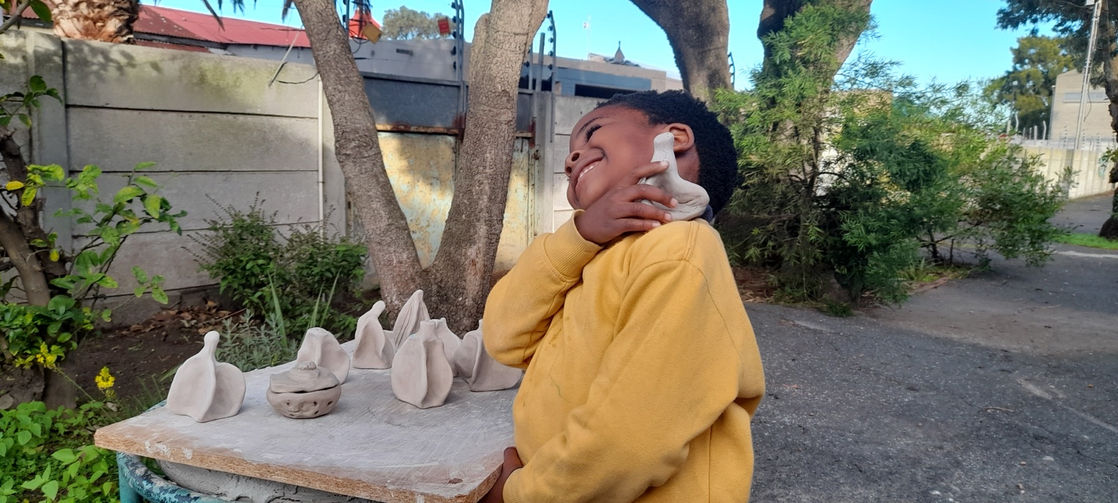 A child outdoors smiles while holding a finished clay sculpture