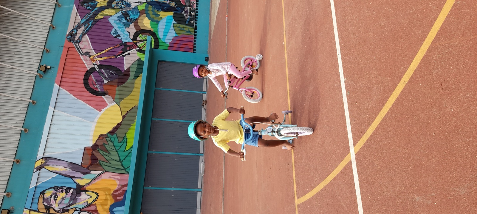 Two children ride bicycles beneath a colorful community mural